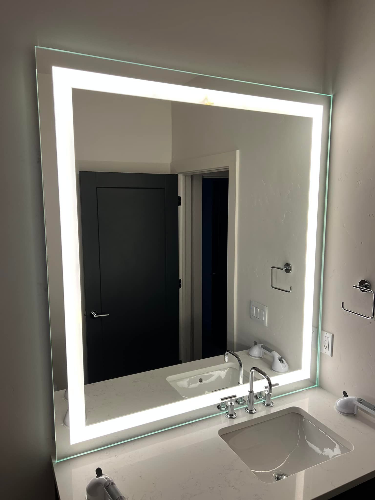 Lit square mirror above a bathroom sink reflecting a dark door.