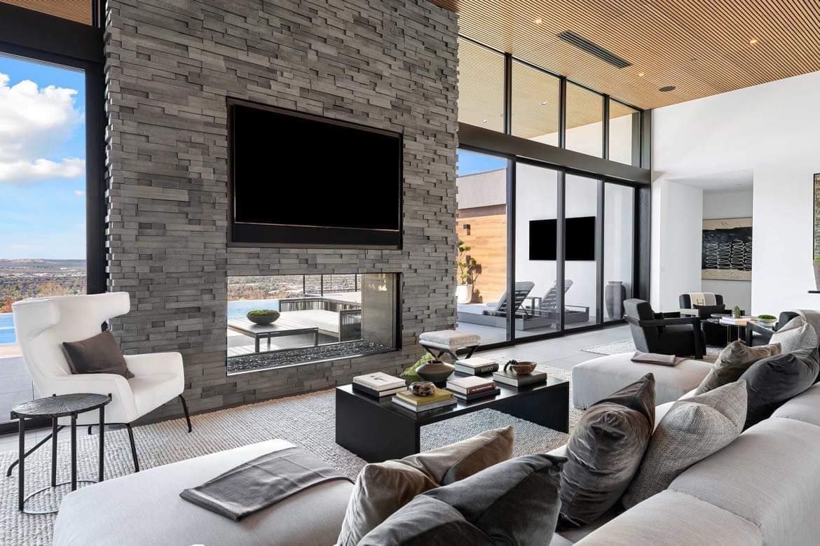 Modern living room with stone fireplace, large TV, white couch, glass walls, and a view.