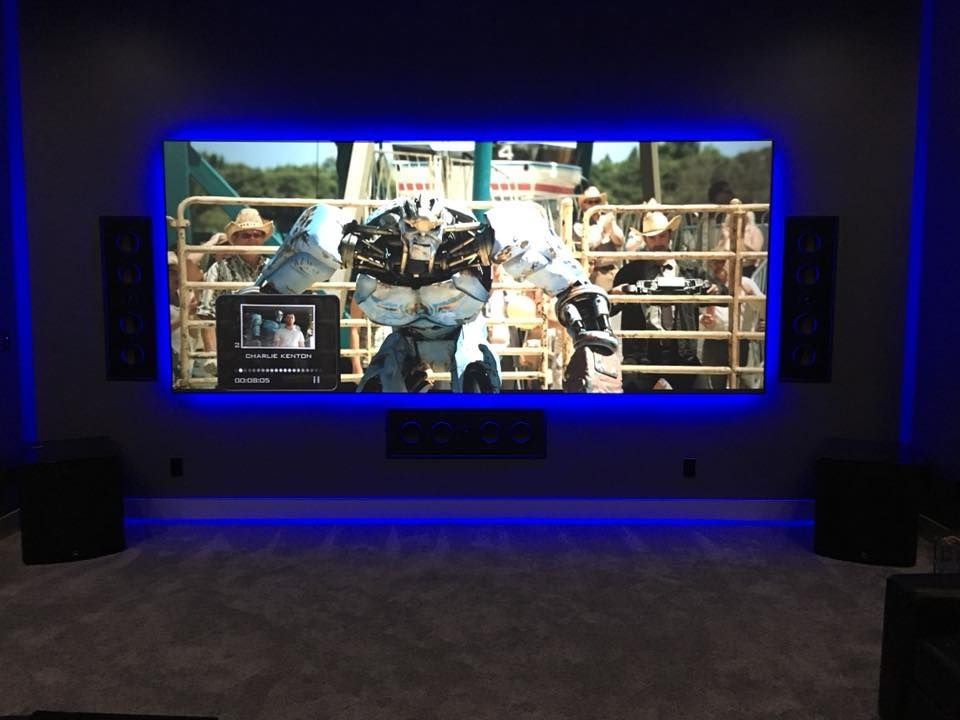 Home theater with large screen, lit blue, playing a movie. Speakers, subwoofer, and gray carpet present.