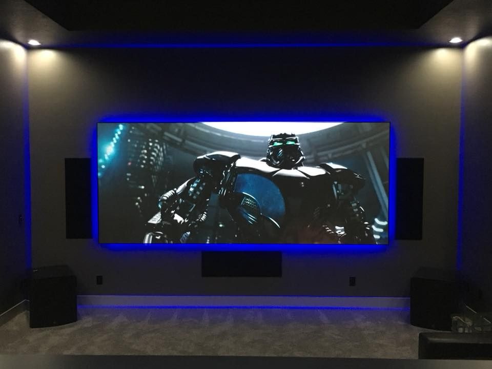 A dark home theater room with a large screen, blue LED border, and speakers; movie scene displayed.