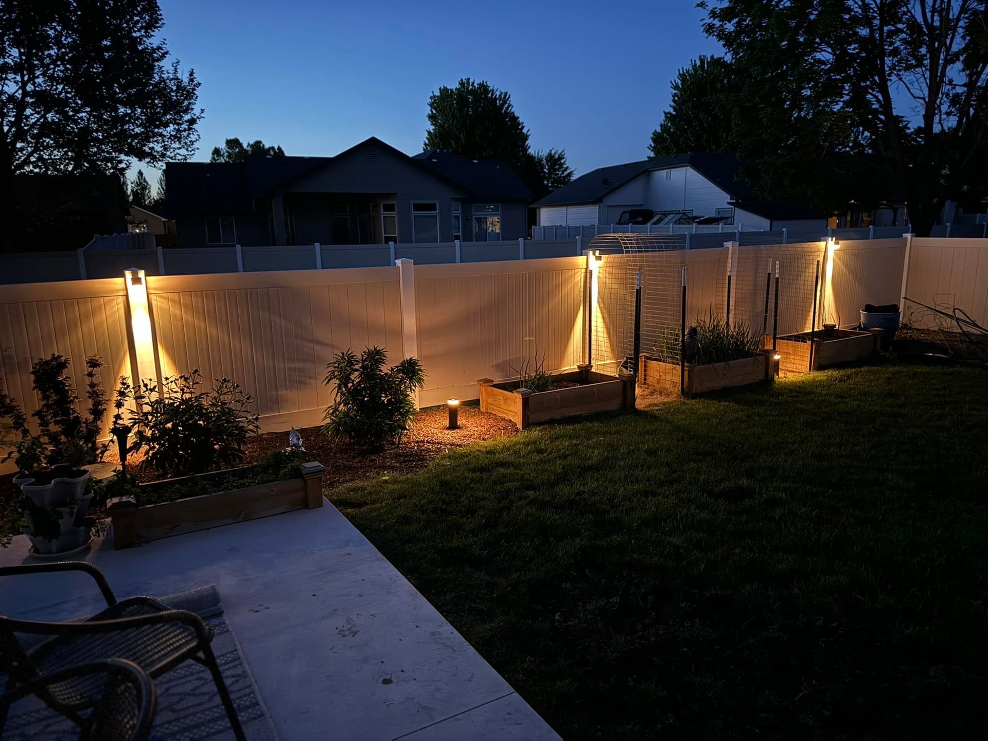 Backyard with illuminated fence, raised garden beds, and two chairs on a concrete patio at dusk.