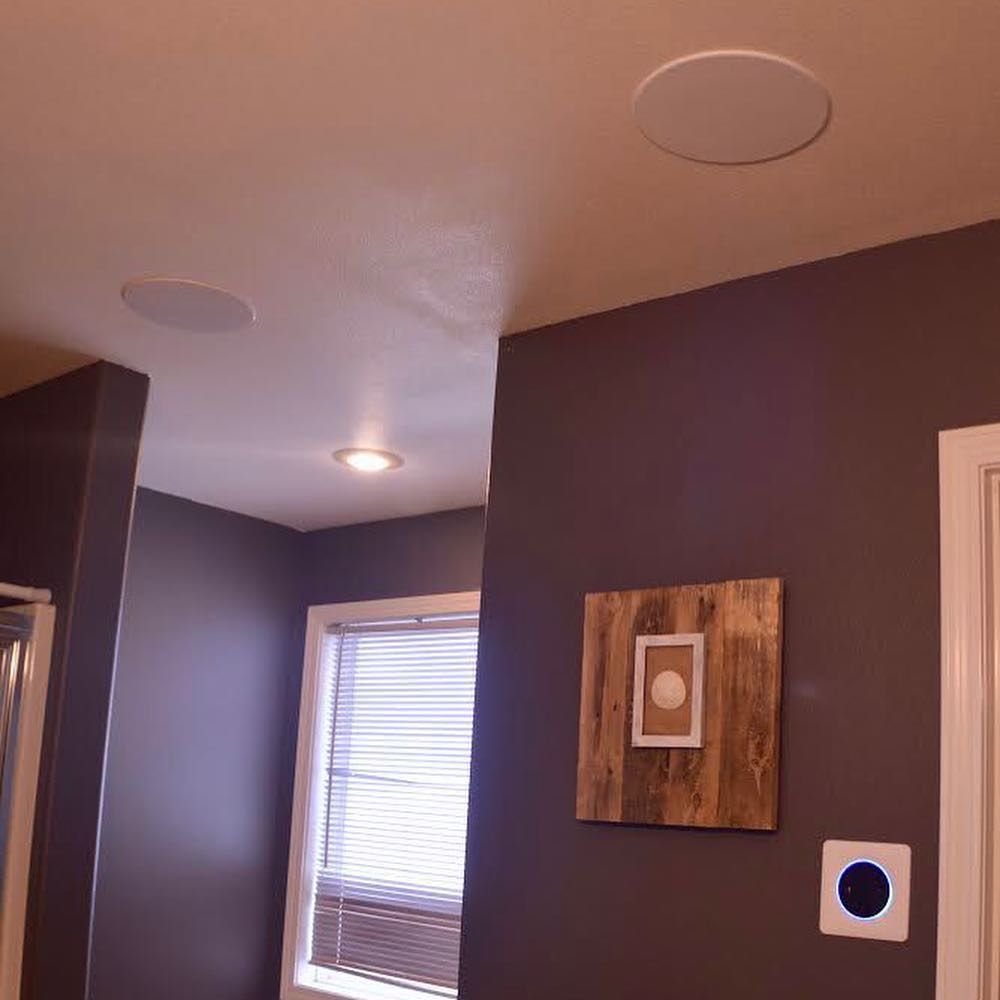 A bathroom ceiling with speakers and recessed lighting. A wooden art piece hangs on the dark purple wall.
