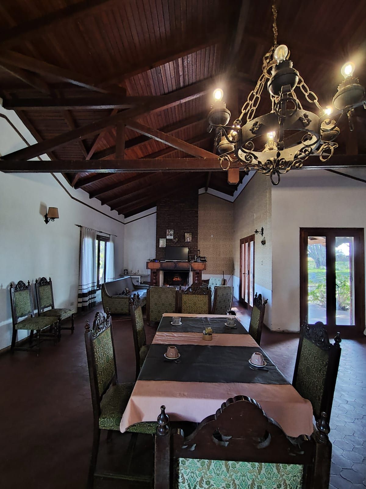 Comedor con mesa larga, sillas ornamentadas, lámpara de araña, chimenea, techo de madera.