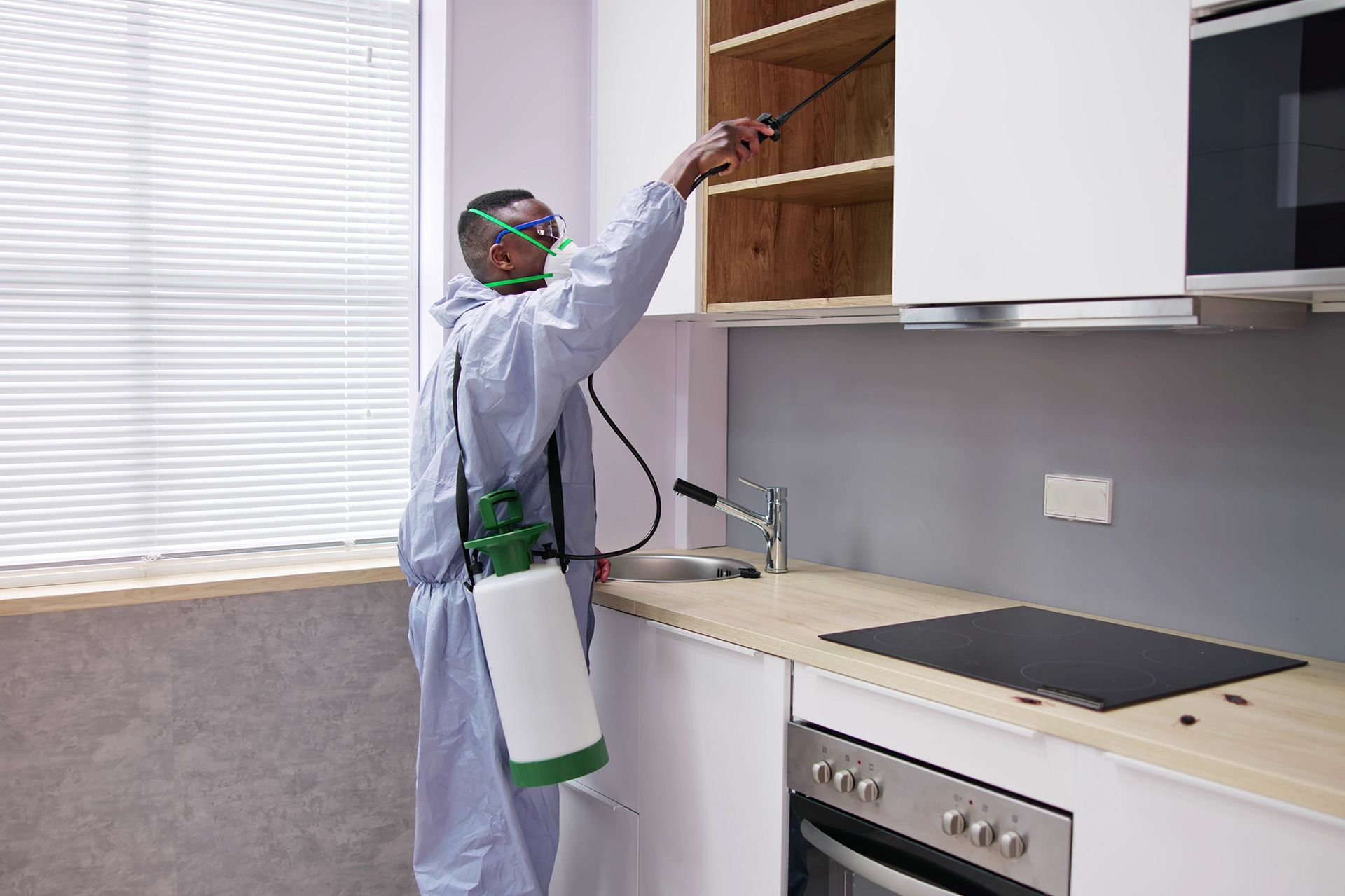 Person in protective suit spraying inside a kitchen cabinet to eliminate pests.