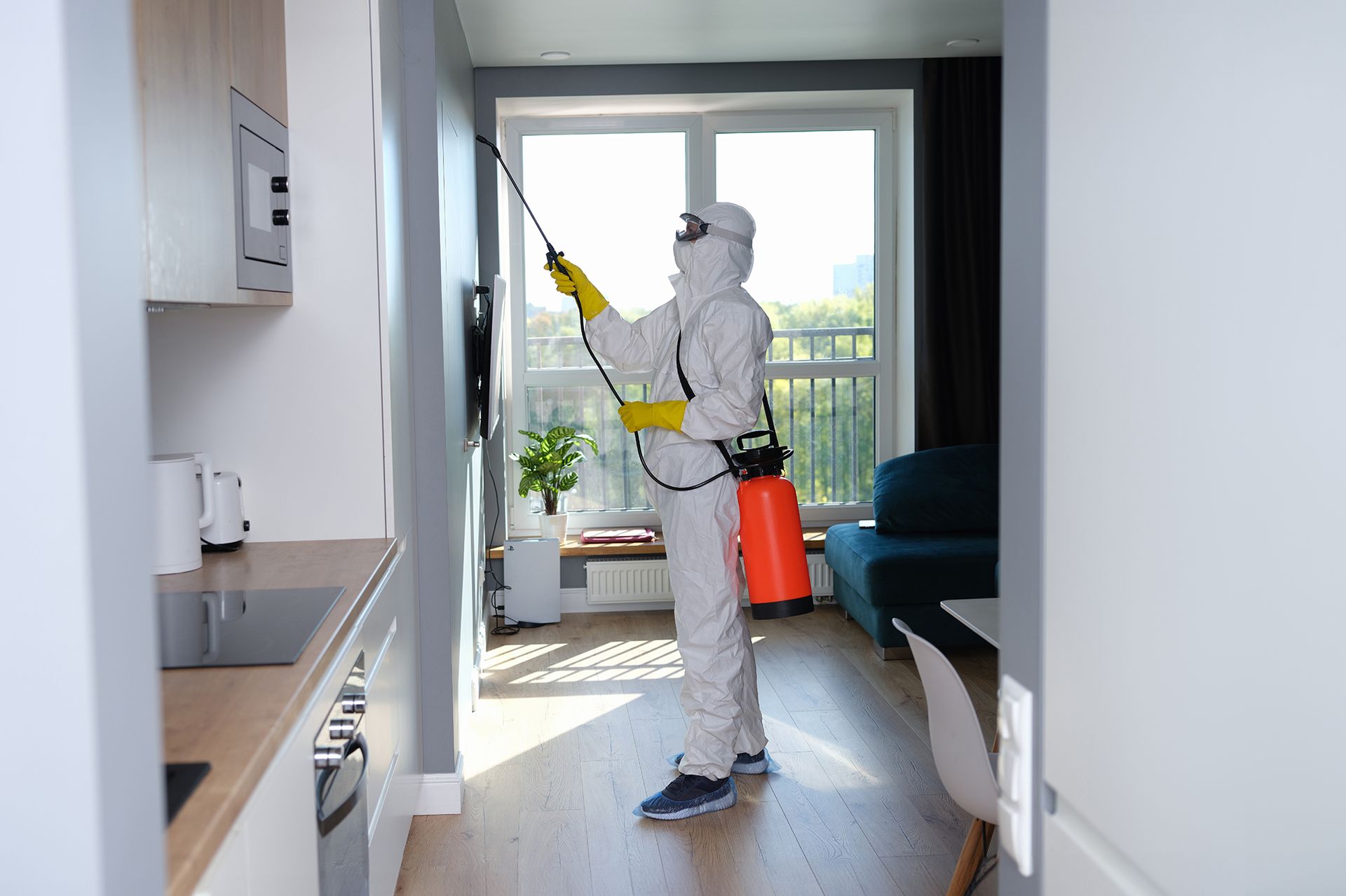 Person in protective suit spraying insecticide in an apartment, next to kitchen and living area.