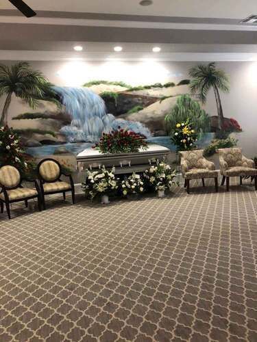 Funeral home interior with casket, floral arrangements, and painted mural of a waterfall.