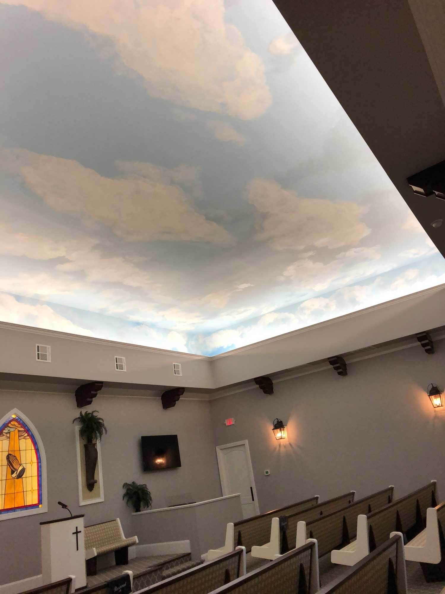 Interior of a chapel with painted sky ceiling, rows of chairs, stained glass, and a small TV.