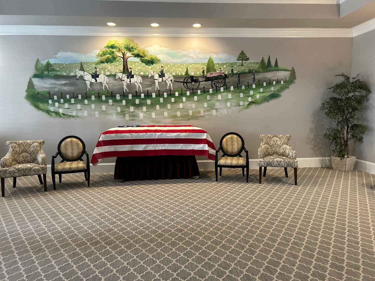 A funeral service with a casket draped in an American flag, four chairs, and a mural of a battlefield.