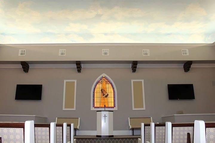 Interior view of a church with a stained glass window, podium, and seating; the ceiling has a cloudy sky mural.