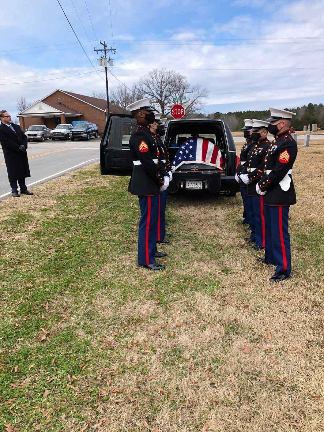 Marines in dress uniform stand at attention near a hearse draped with an American flag.