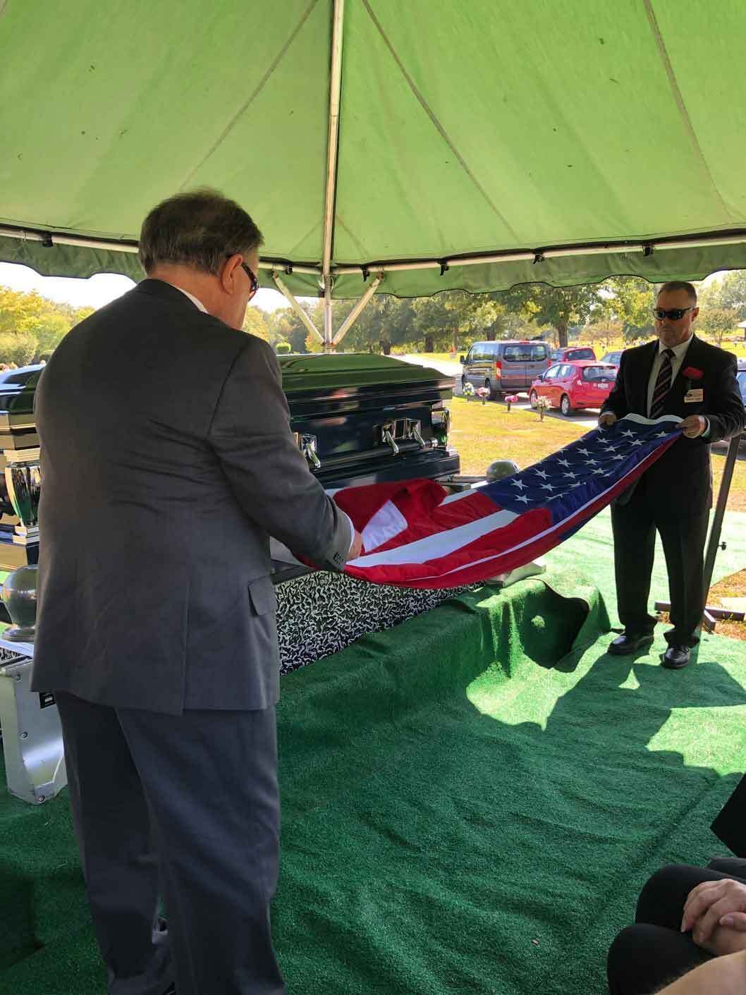 Men folding an American flag over a casket at an outdoor funeral under a tent.
