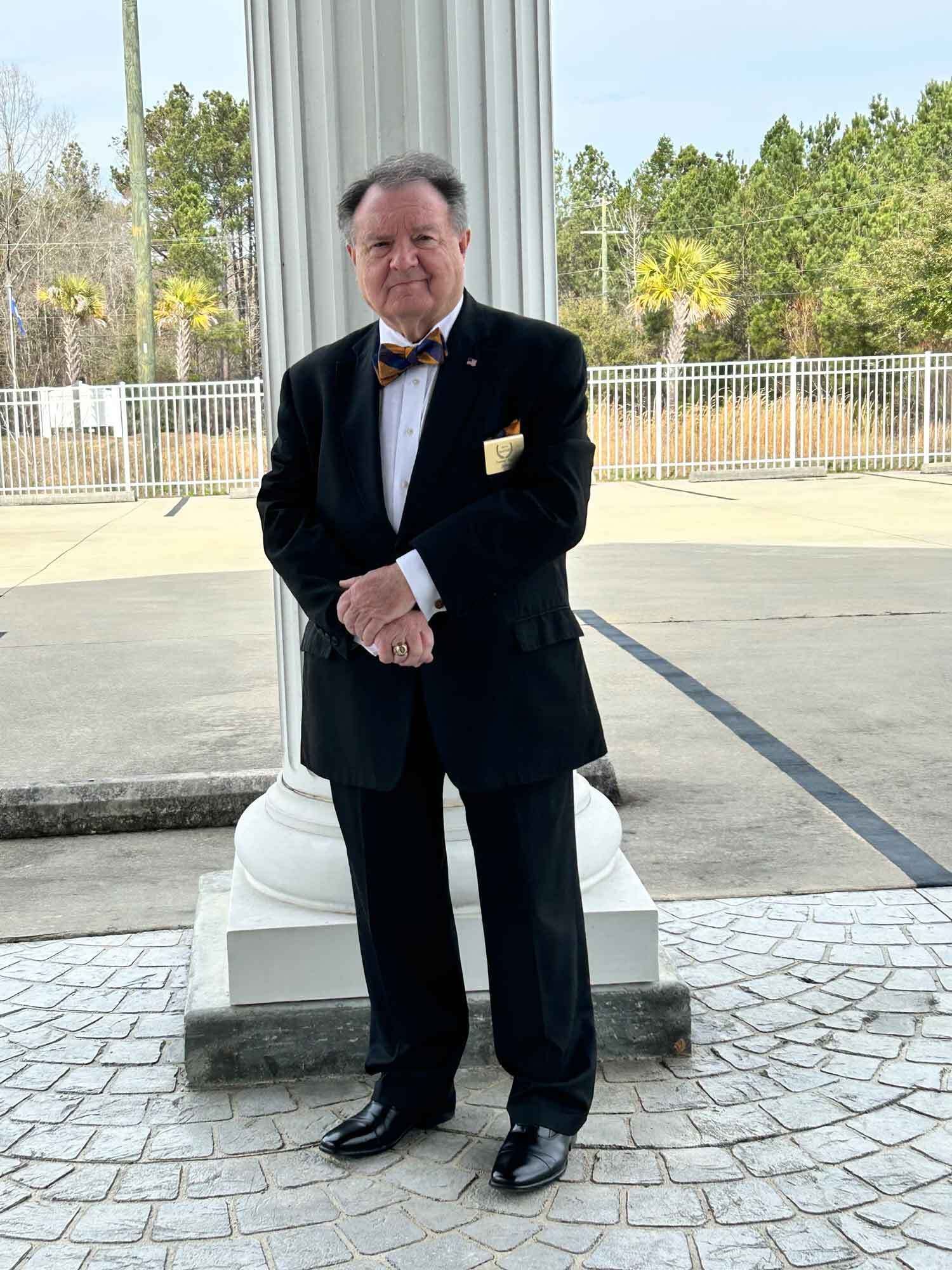 Man in black tuxedo stands beside a white column, hands clasped in front. Outdoor setting.