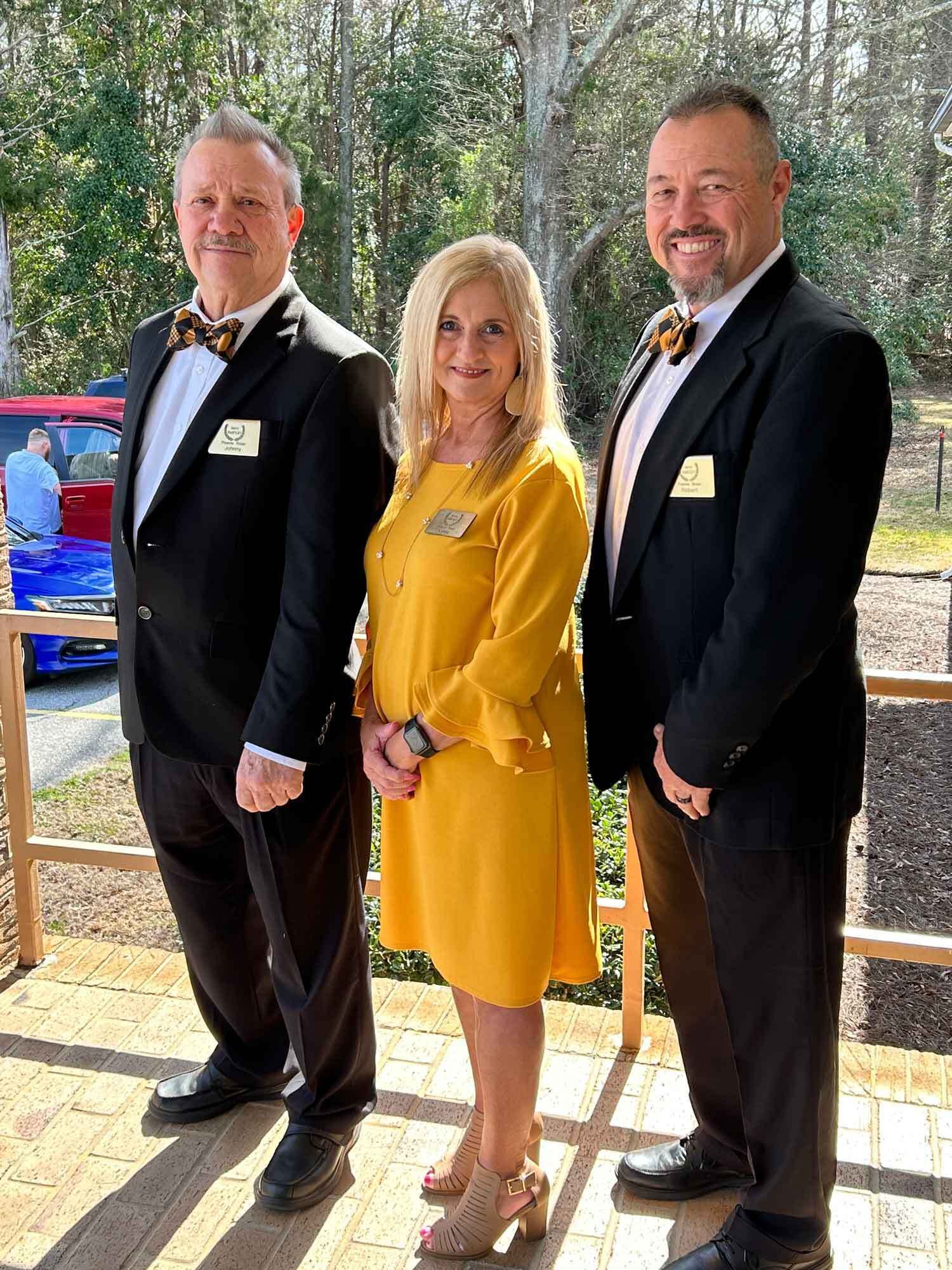 Three people pose outdoors: woman in yellow dress flanked by men in black suits with bow ties.