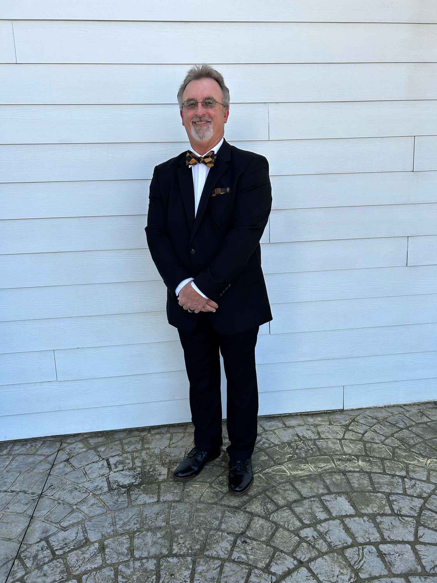 Man in black suit and patterned bow tie stands in front of a white wall.