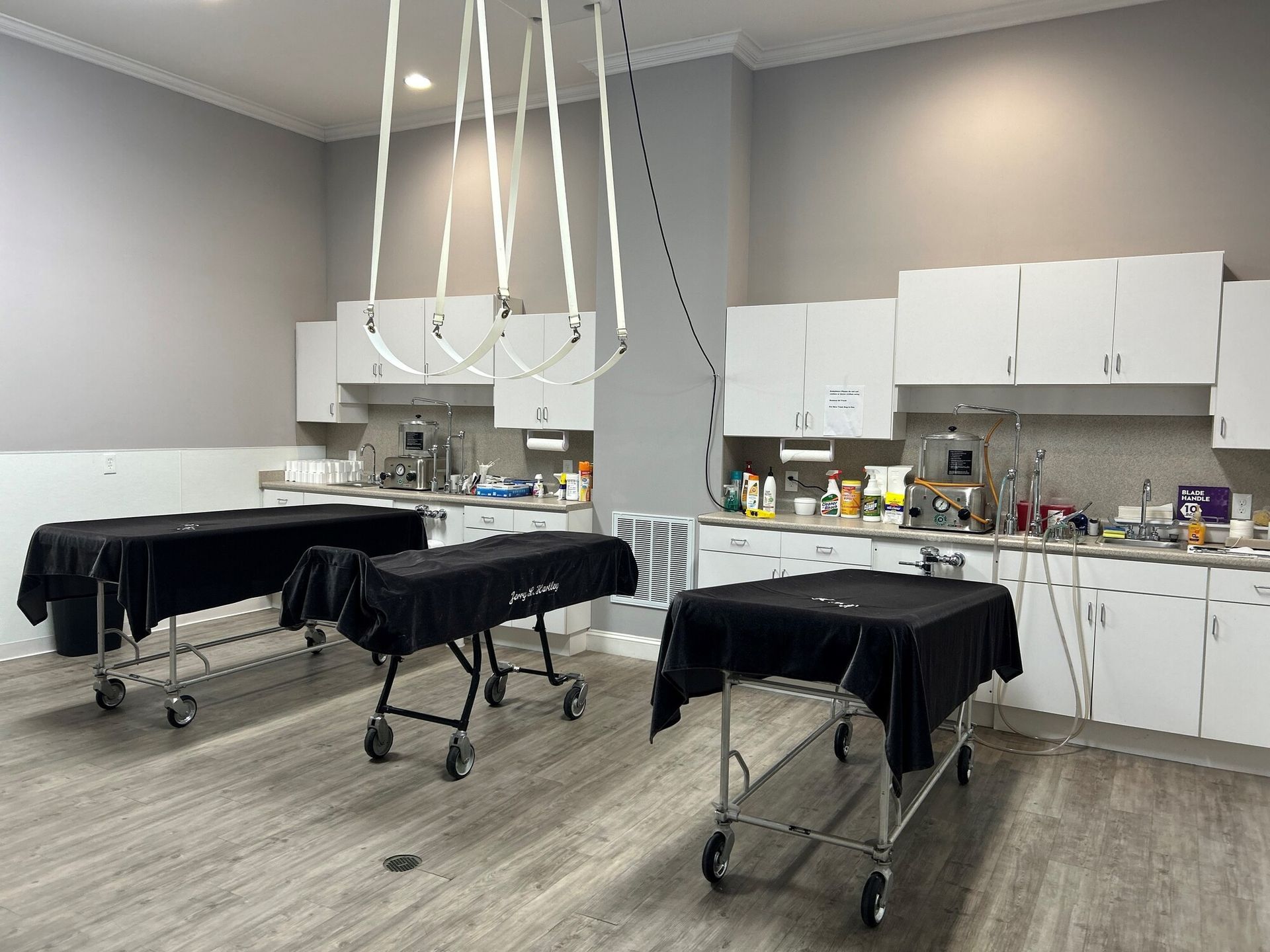 A mortuary with three gurneys covered in black cloth. White cabinets and gray walls.