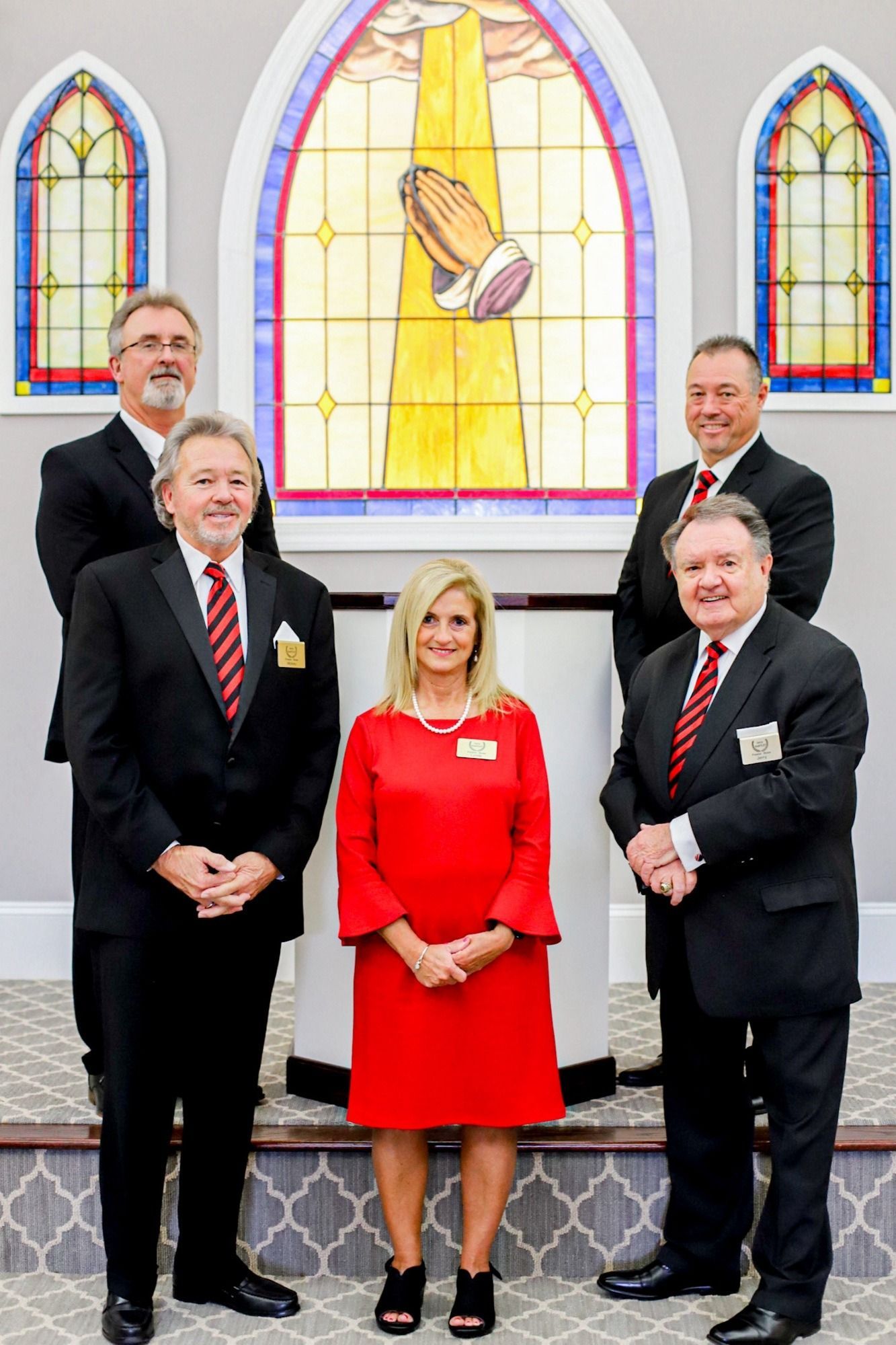 Group of six formally dressed people posing in front of a stained-glass window in a church.