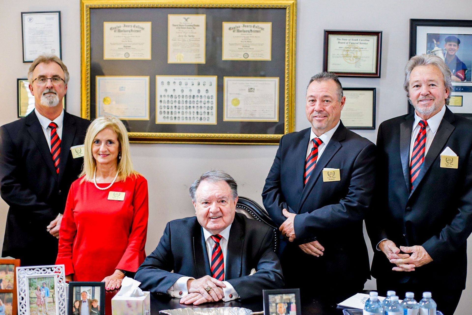 Group of formally dressed people posing together, indoors, by a desk and framed documents.