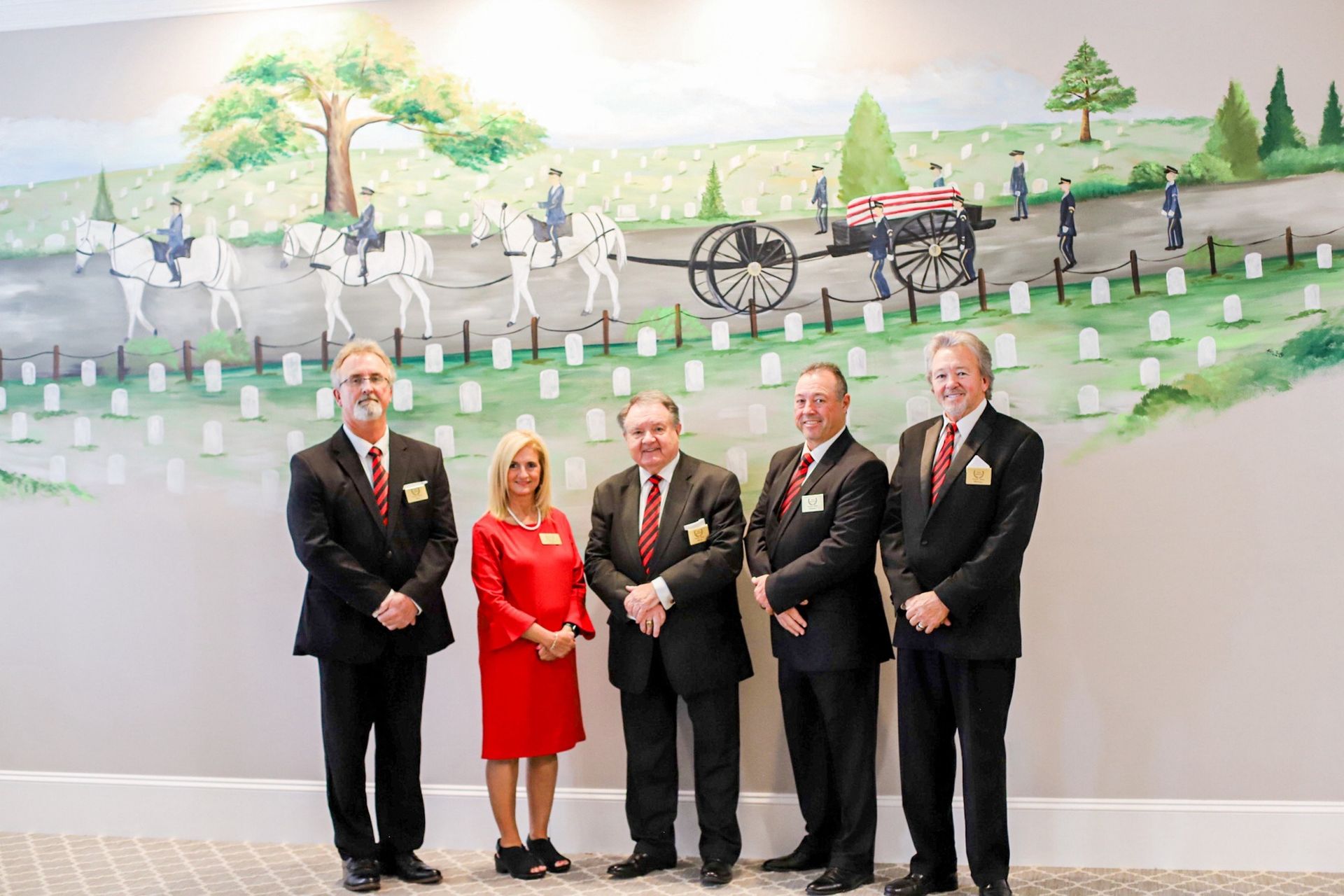 Five people standing in front of a cemetery mural. Men in suits, one woman in red dress.