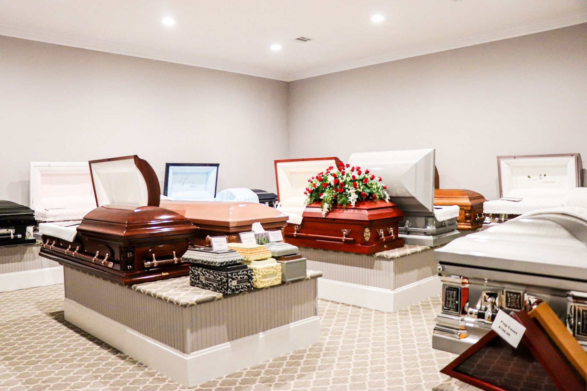 A room displays various open and closed caskets on platforms, showcasing funeral home options.