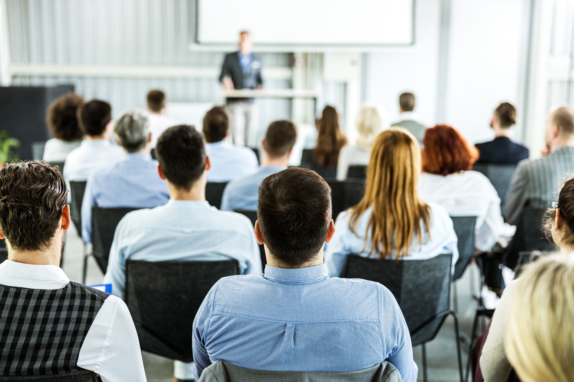 A business presentation in a modern conference room. A man speaks from a podium to a seated audience.