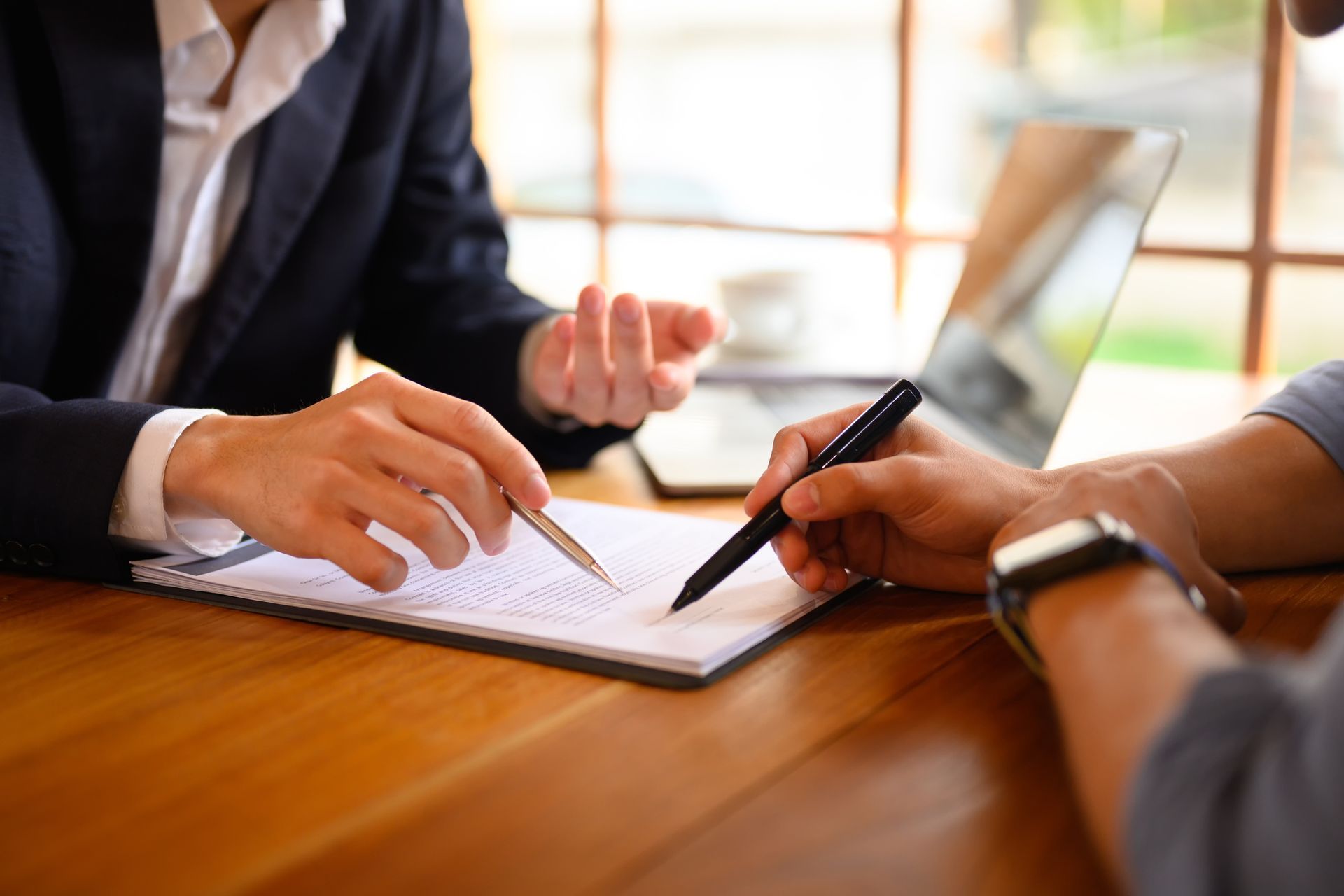 Two people at a wooden table review paperwork. One points with a pen, the other prepares to sign a document.