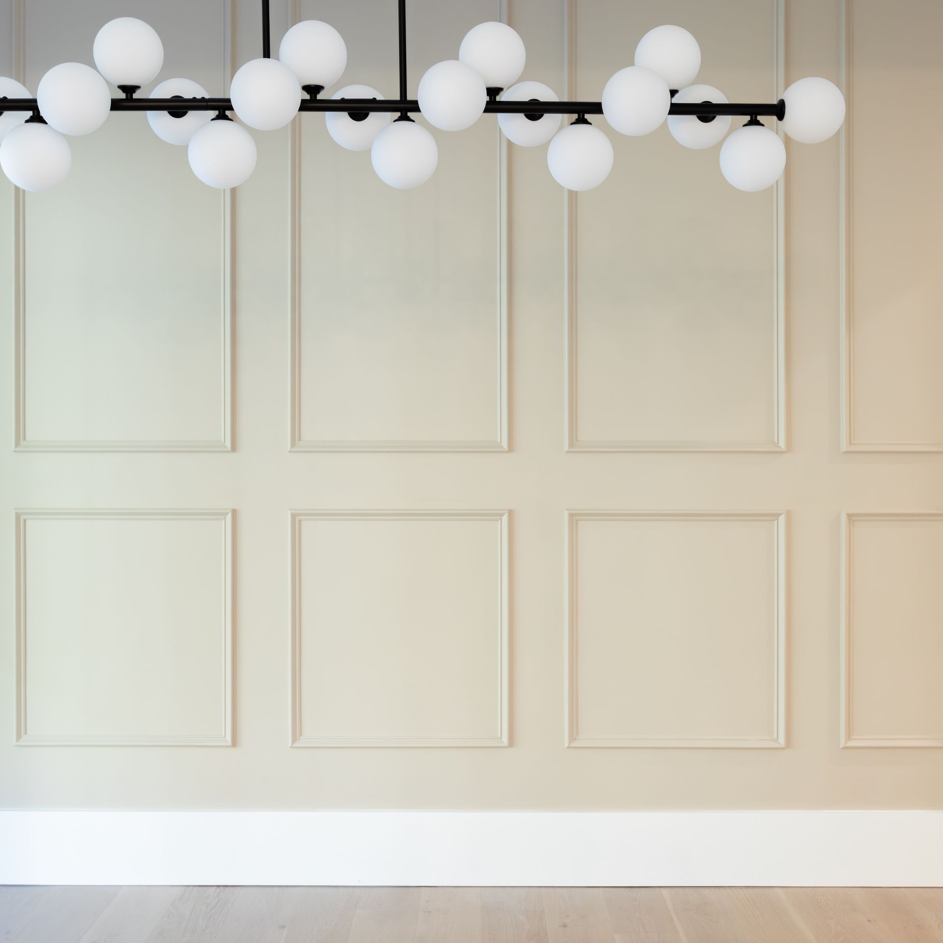 A modern chandelier with white globes hangs above a beige paneled wall and white baseboard.