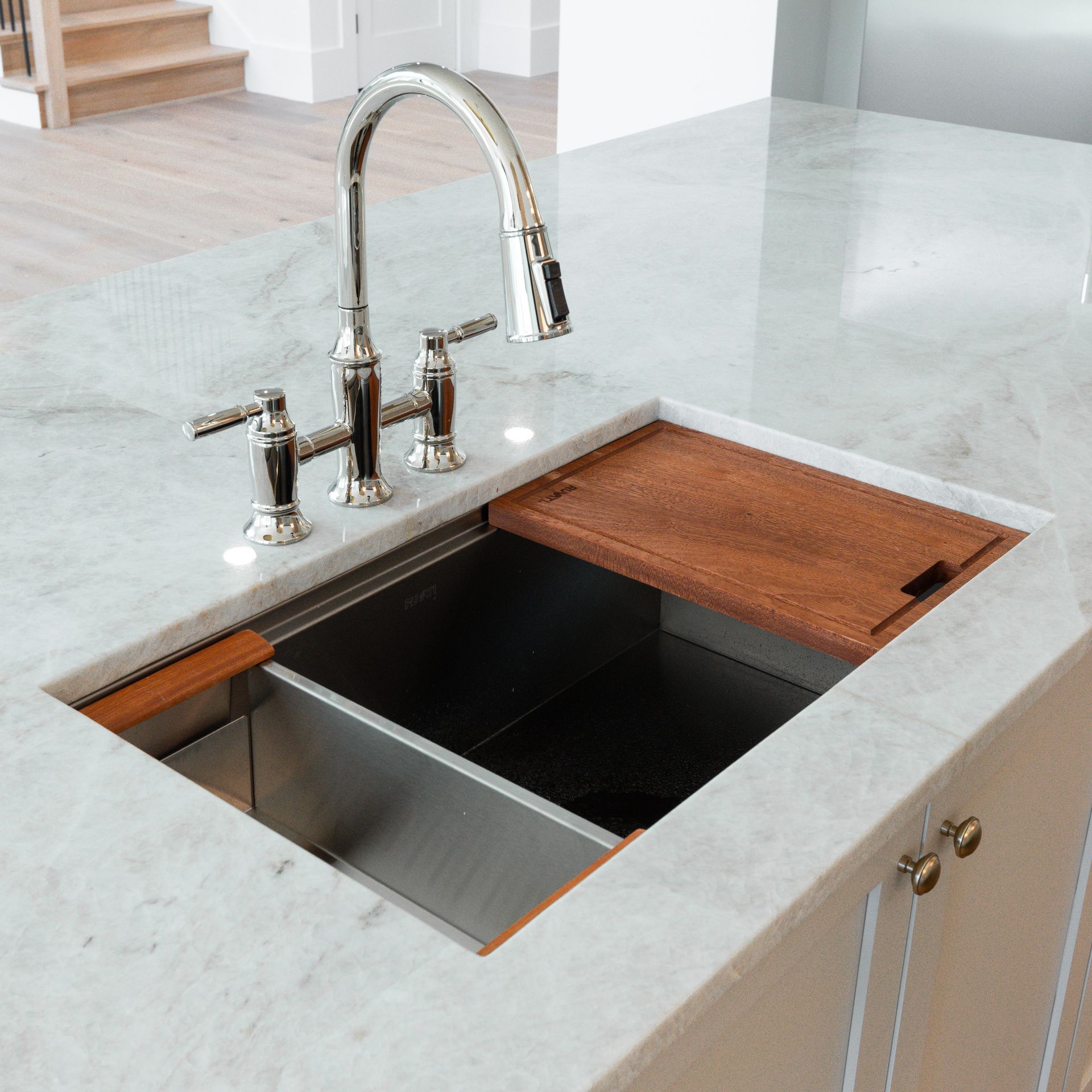 Stainless steel kitchen sink with wooden cutting board and chrome faucet on marble countertop.