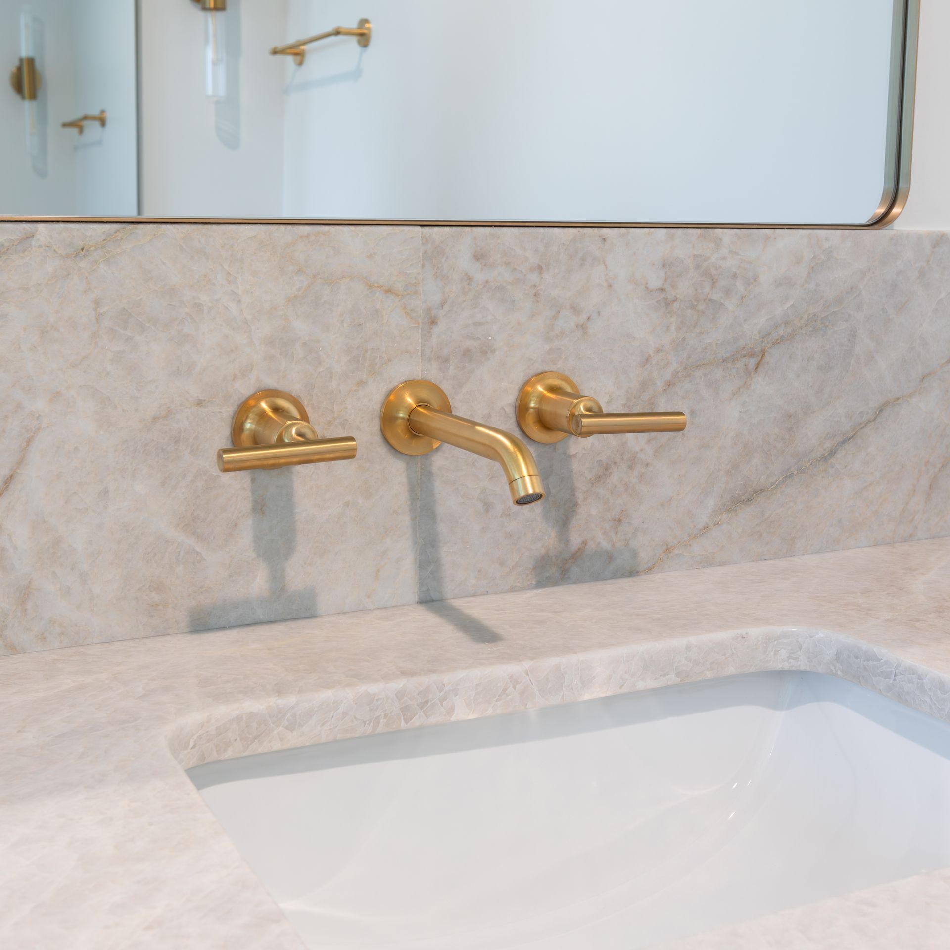 Gold-toned wall-mounted faucet and handles above a white sink in a bathroom with a marble countertop.