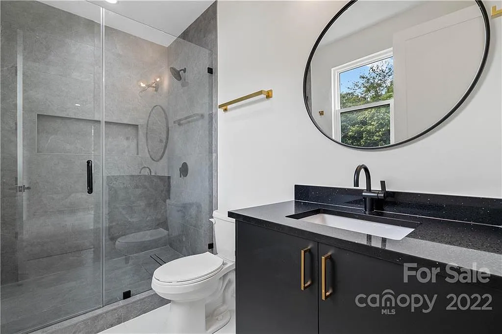 Modern bathroom with a glass shower, vanity, and toilet. Black vanity with gold hardware and an oval mirror.