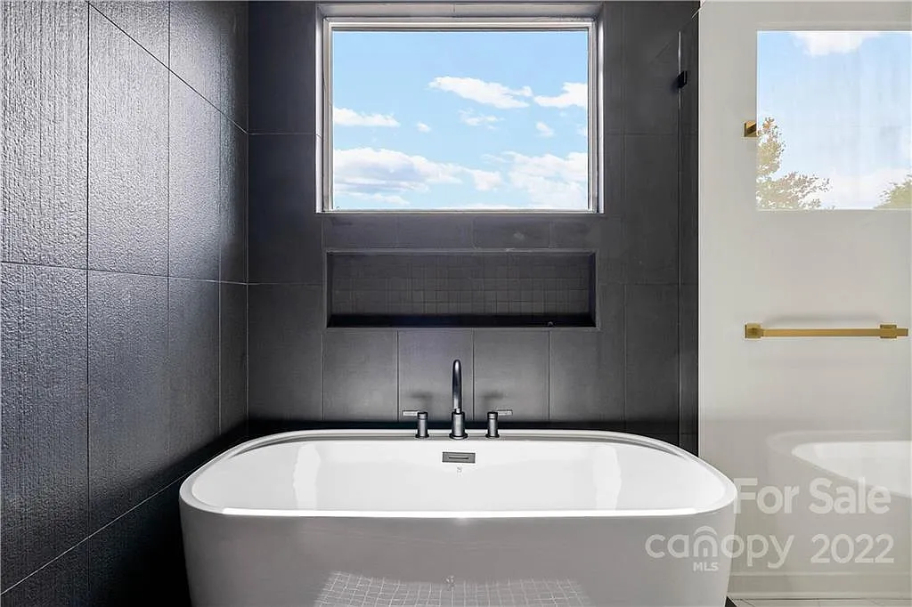 Modern bathroom with a white tub, gray tile, and a window with a sky view.