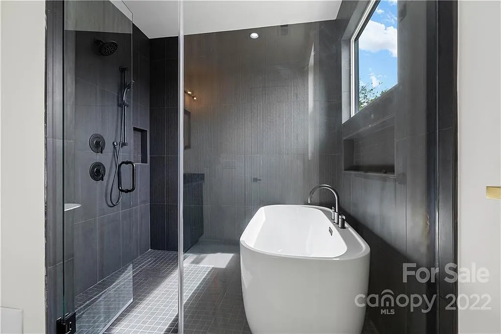 Modern bathroom with glass shower, soaking tub, and window. Dark gray walls, white tub.