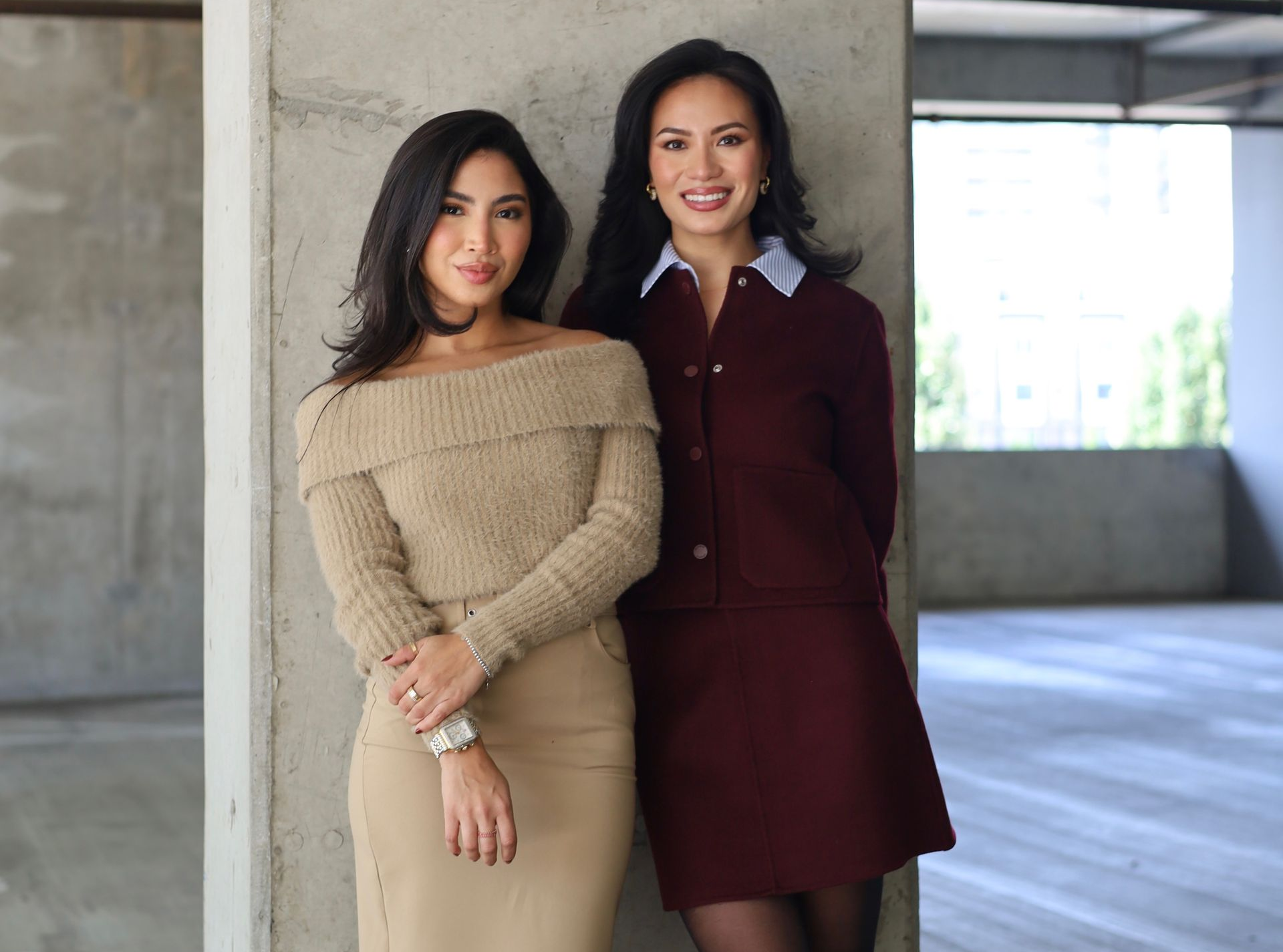 Two women, one in beige off-shoulder sweater, the other in a burgundy suit, lean against a concrete column.