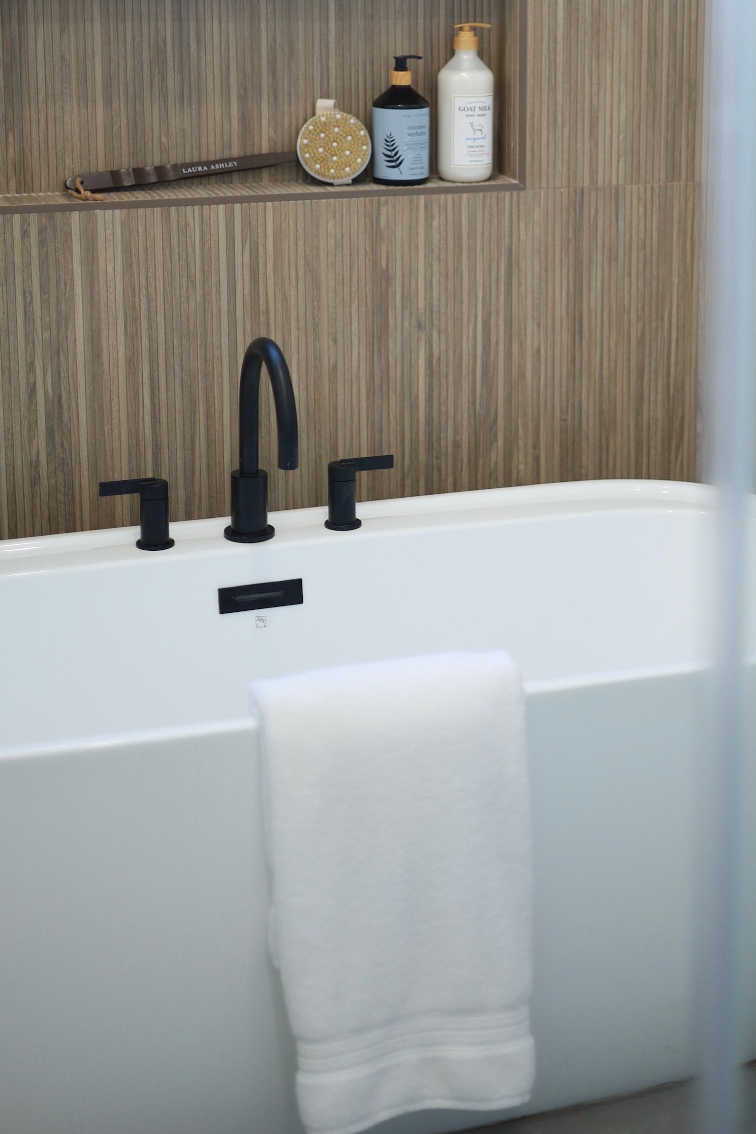 A modern bathroom with a white tub, black faucet, and wooden-look tiled wall. A white towel hangs on the tub.