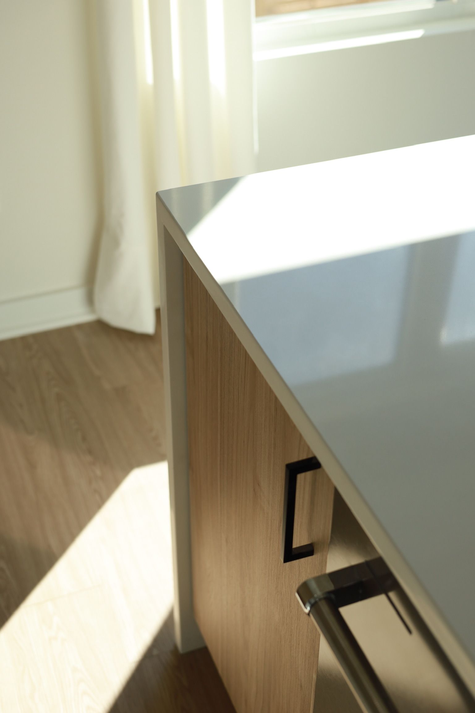 Close-up of a kitchen island with a white countertop, wood-grain panel, and black handle. Natural light shines.