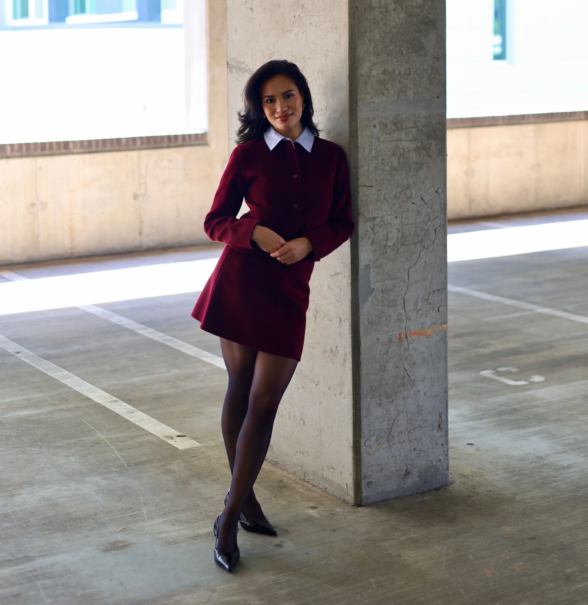 Woman in burgundy suit and black tights leans against a concrete pillar in a parking garage.