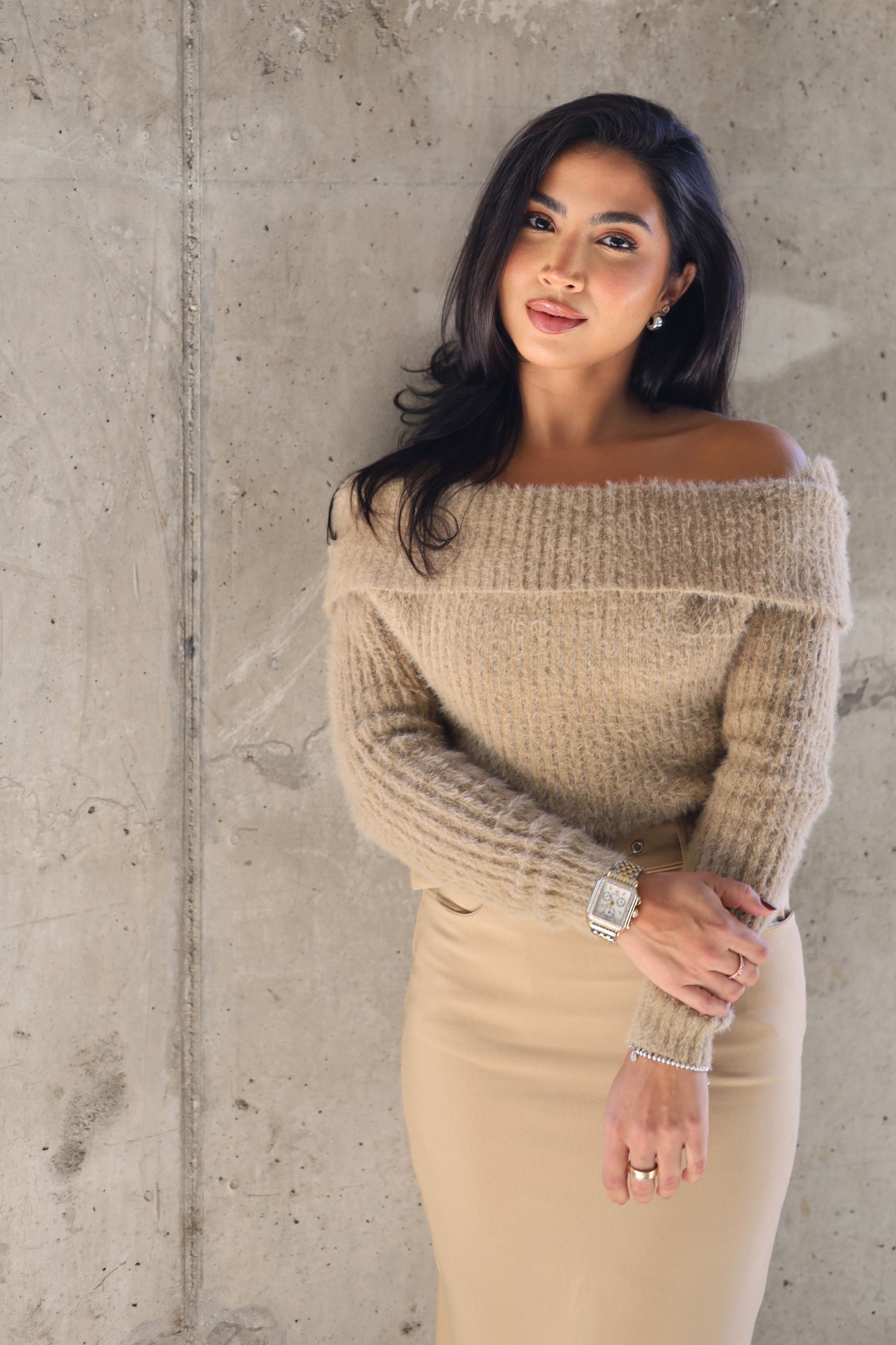 Woman in beige off-the-shoulder sweater and skirt, leaning against a gray concrete wall, looking at the camera.