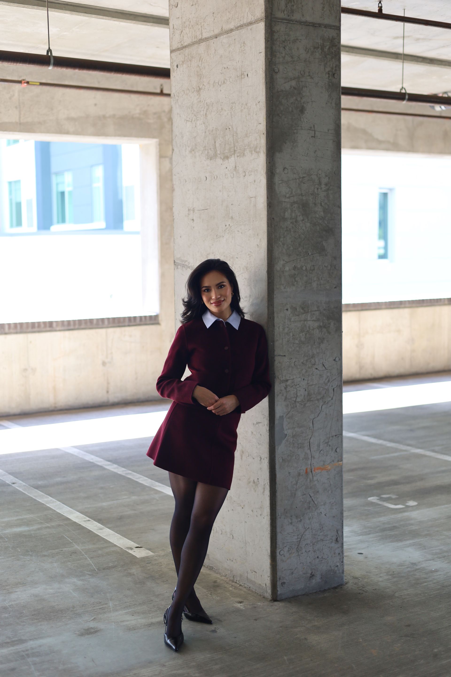 Woman in a maroon dress and white collar leans against a concrete pillar in a parking garage.