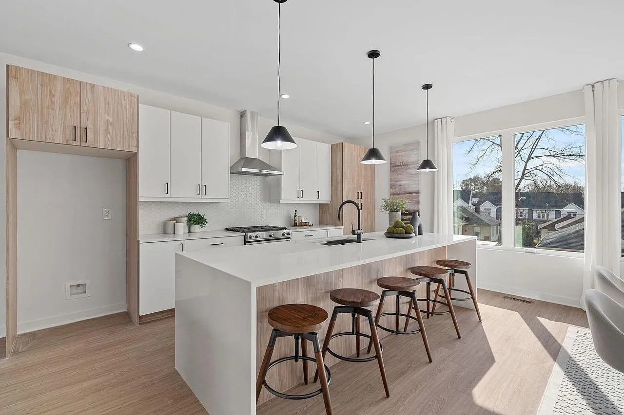 Modern kitchen with white cabinets, wooden island, and stools; large windows with a view.