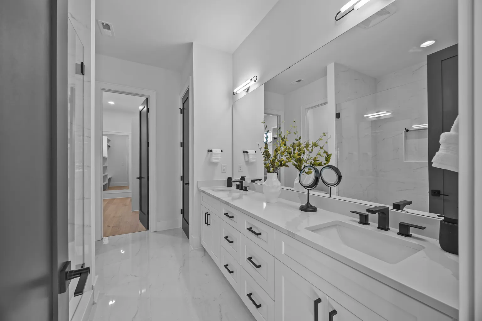 White bathroom with double sinks, large mirror, and light fixtures. Black accents, doorway visible.