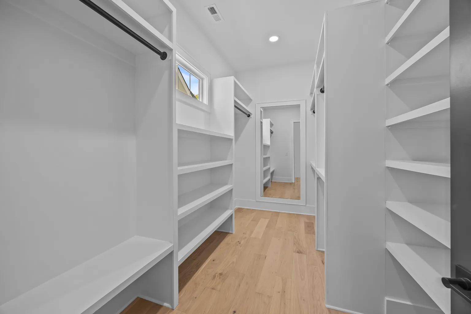 Walk-in closet with white shelving, light wood floor, and a mirror.