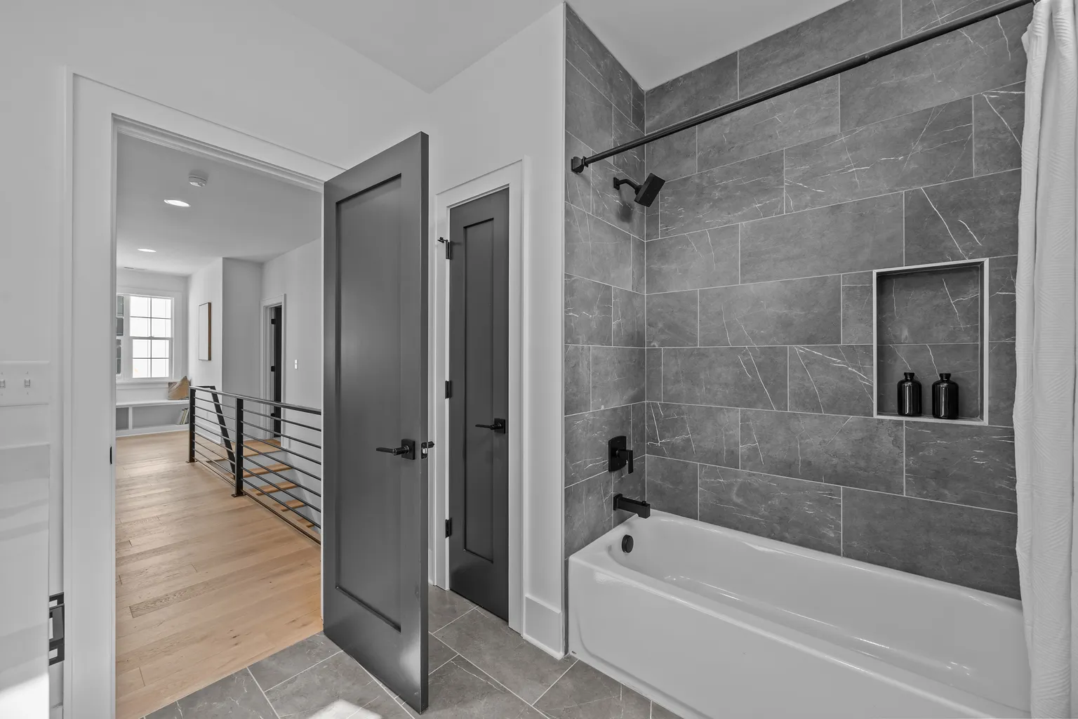 Bathroom with gray tiled shower, white bathtub, and a hallway with hardwood floors and black railing.