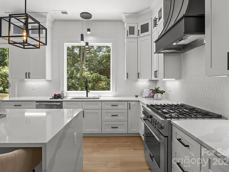 Bright white kitchen with a gas stove, cabinets, and a large window overlooking greenery.