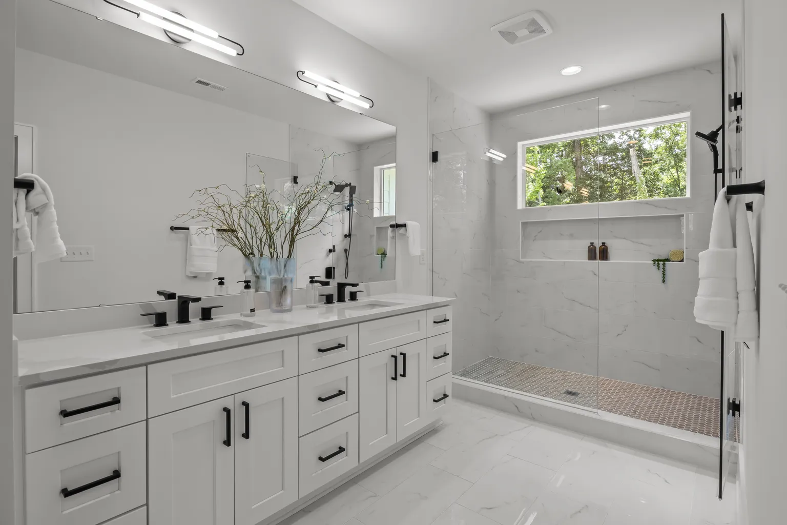 White bathroom with a double vanity, glass shower, and natural light from a window.