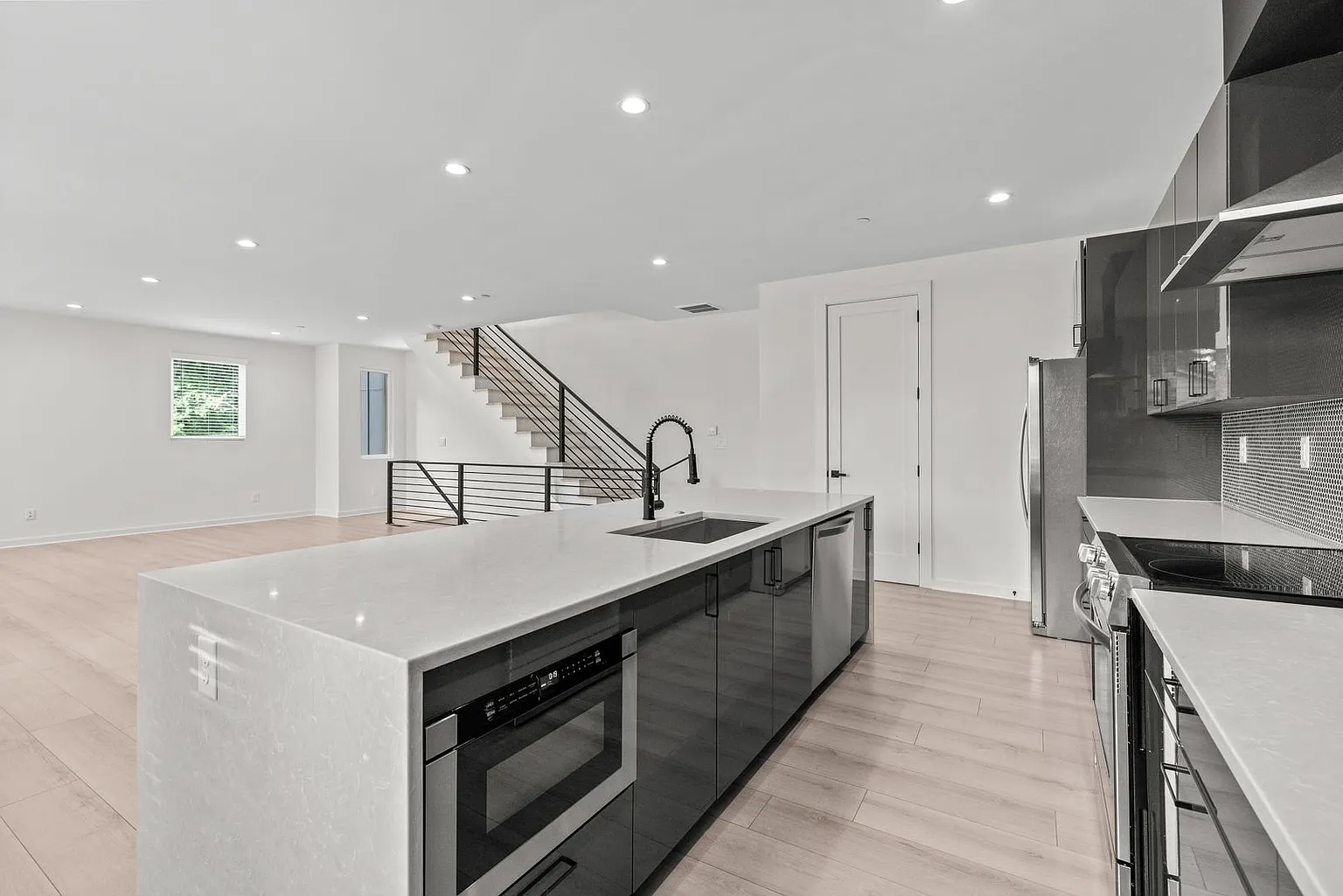 Modern kitchen with island, stainless appliances, light wood floors, and a staircase in the background.