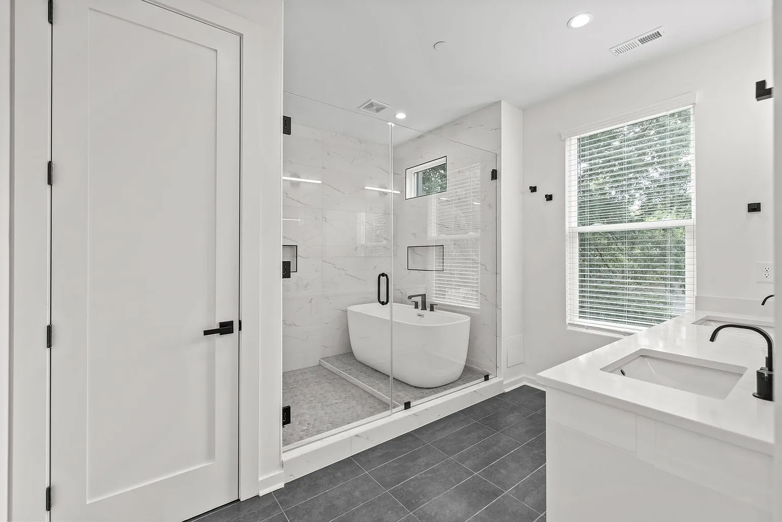 Modern white bathroom with a glass-door shower, freestanding tub, double vanity, and a window.