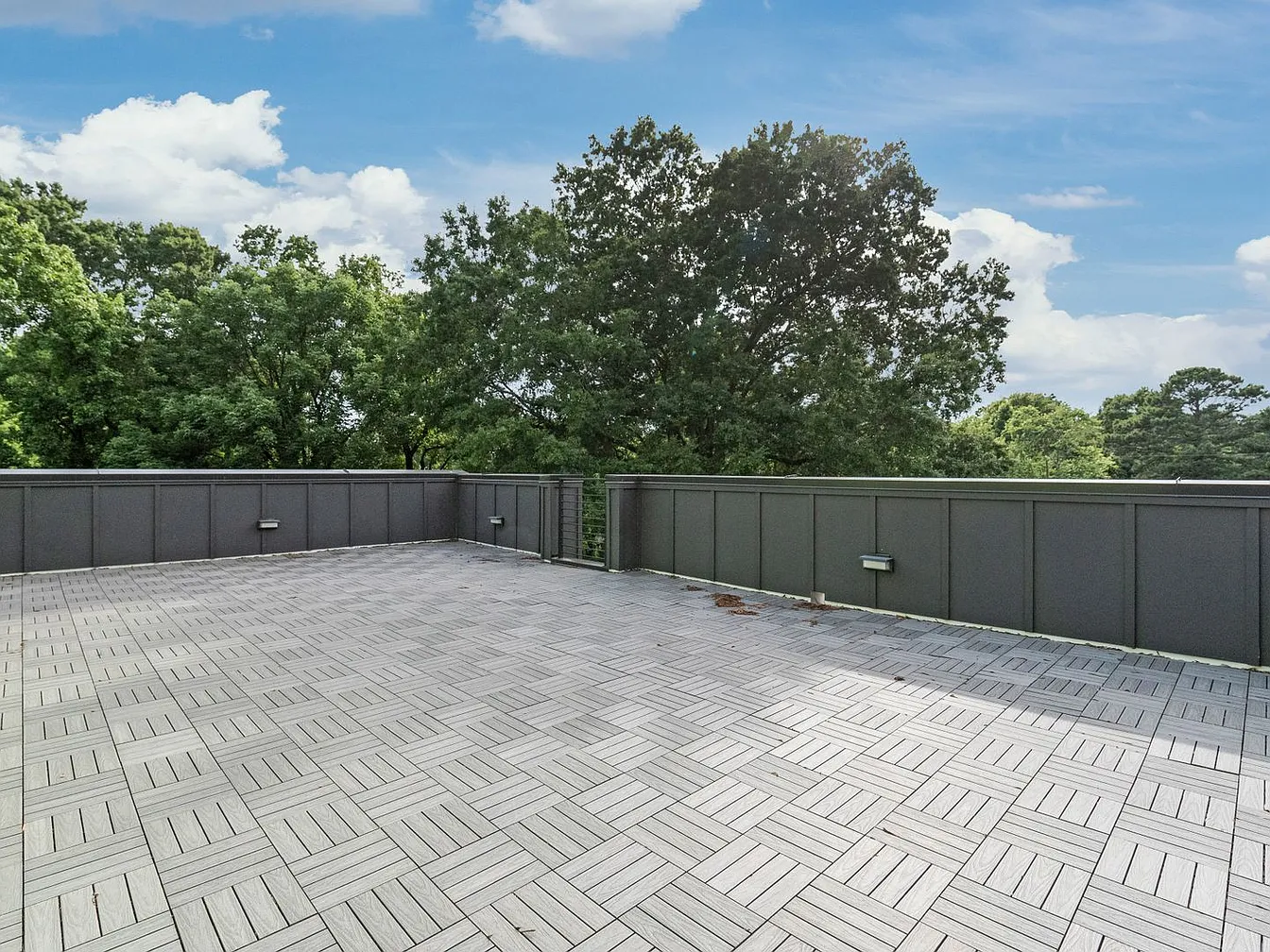 A gray tiled rooftop patio with a dark border and trees in the background under a blue sky with clouds.
