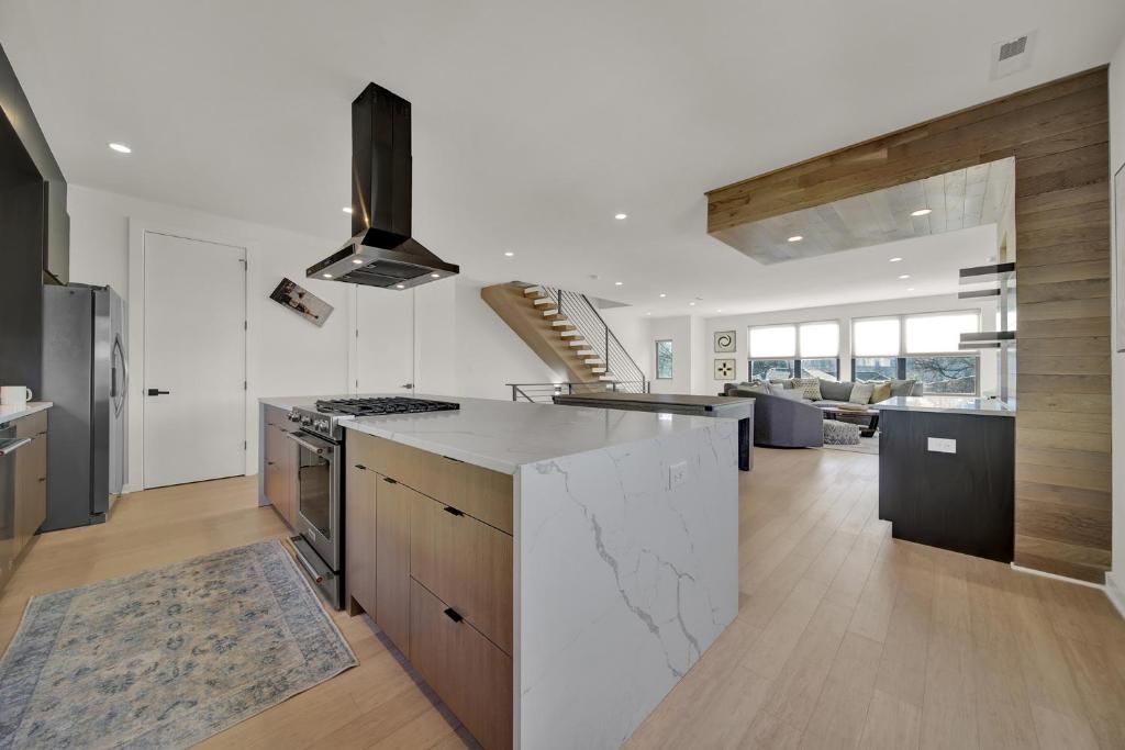 Modern kitchen with island, wooden cabinets, and marble countertop. Open to living area, stairs in background.