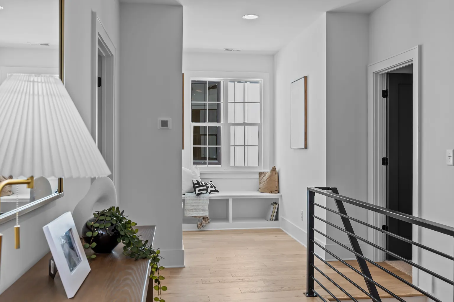 Hallway with light wood floors, bench under a window, black door, and metal railing.