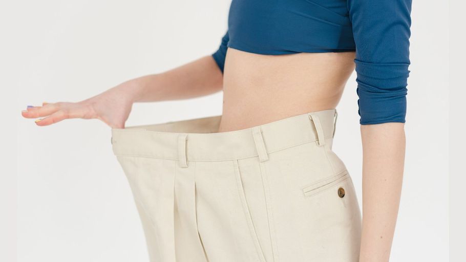 Woman in blue top holding out loose beige pants, showing weight loss.
