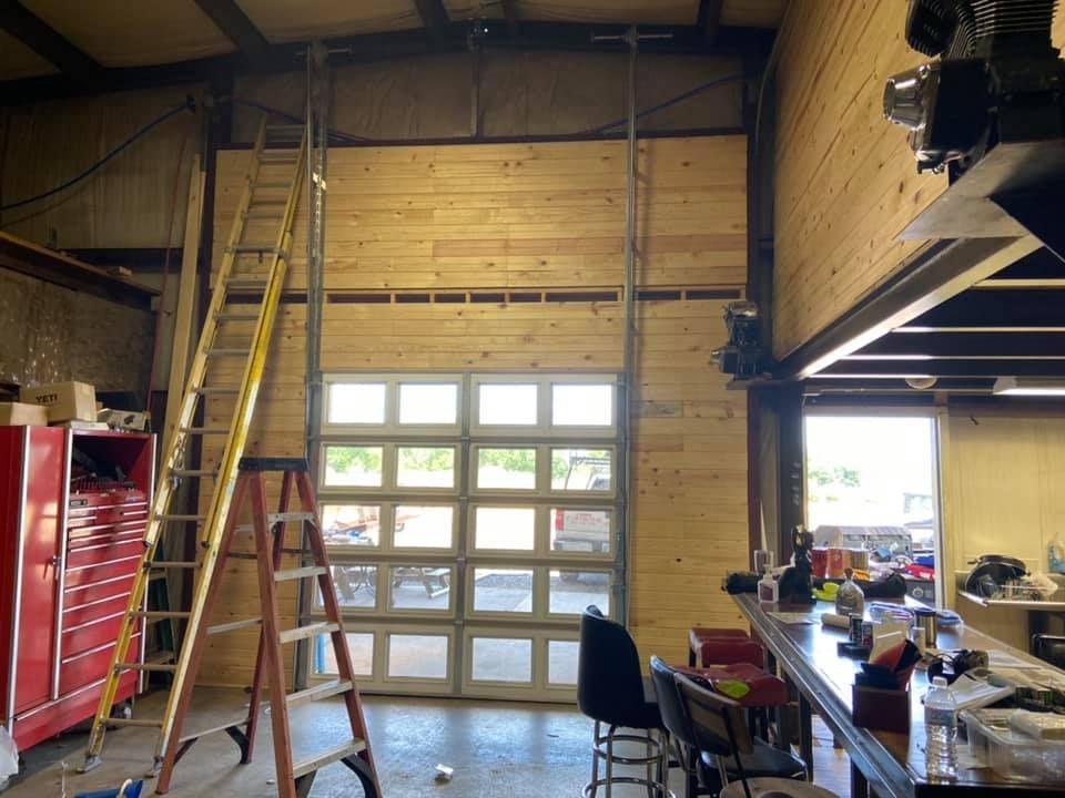 Interior of a workshop with a partially installed glass garage door and wood paneling. A ladder is present.